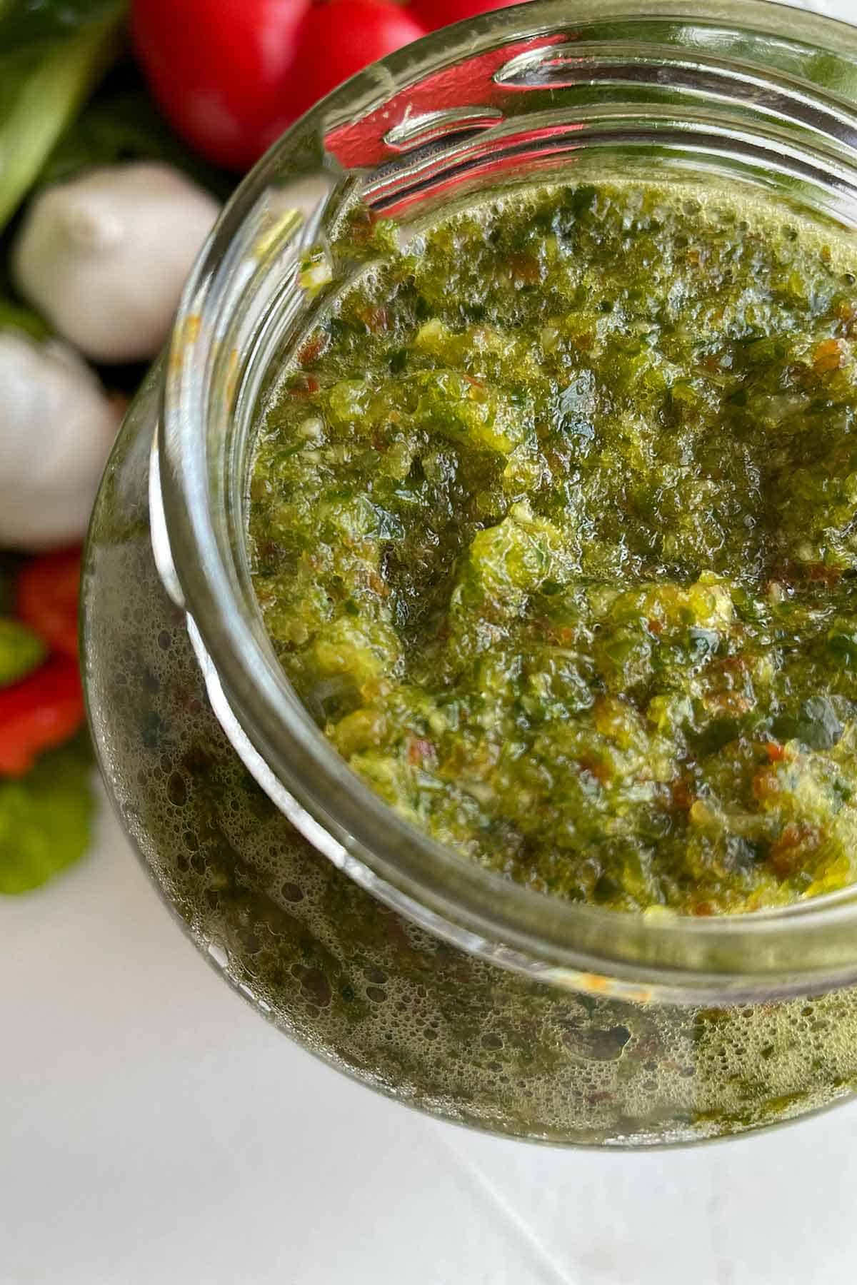 a glass jar full of homemade sofrito with some vegetables in the background.