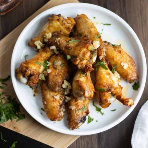 white plate with garlic butter chicken wings on top of a cutting board with a some parsley and knife on the side.