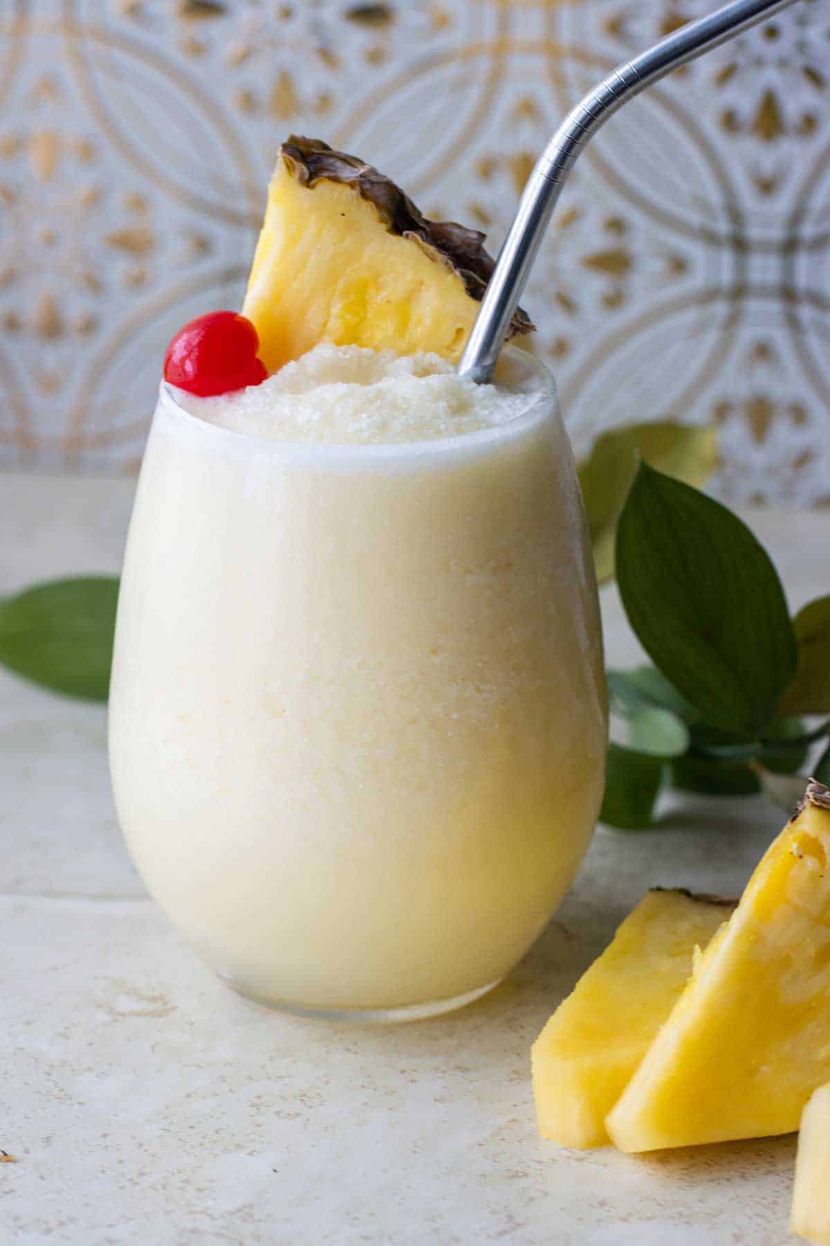 vaso de una piña colada sin alcohol decorada con una cereza y un pedazo de piña. Al lado tiene varios pedazos de piña y unas hojas verdes.