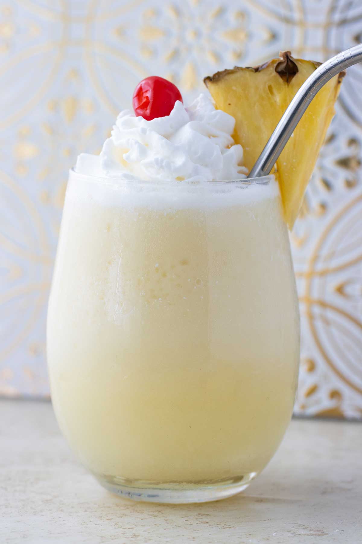 a close up image of a glass cup with pina colada topped with whipped cream, a cherry and a slice of pineapple on the side.