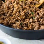 close up of a cast iron with puerto rican picadillo with a wood spoon. Next to it it has a small white dish with olives and parsley.