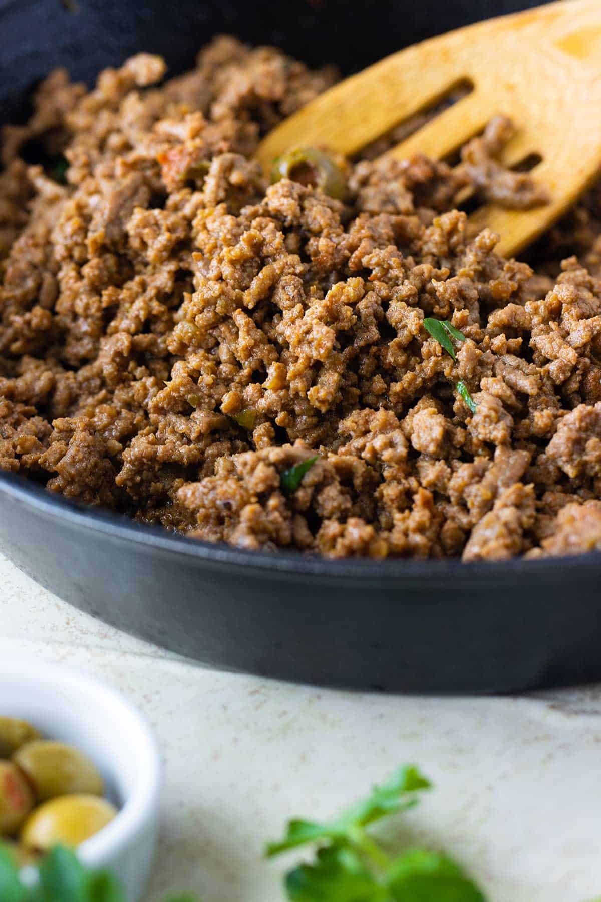 un sarten negro lleno de picadillo de carne molida hecha al estilo puertorriqueno con un cucharon de madera y un envase con aceitunas de lado.