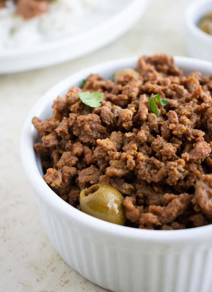 close up of a small white dish with puerto rican picadillo with olives and a plate of food in the background.