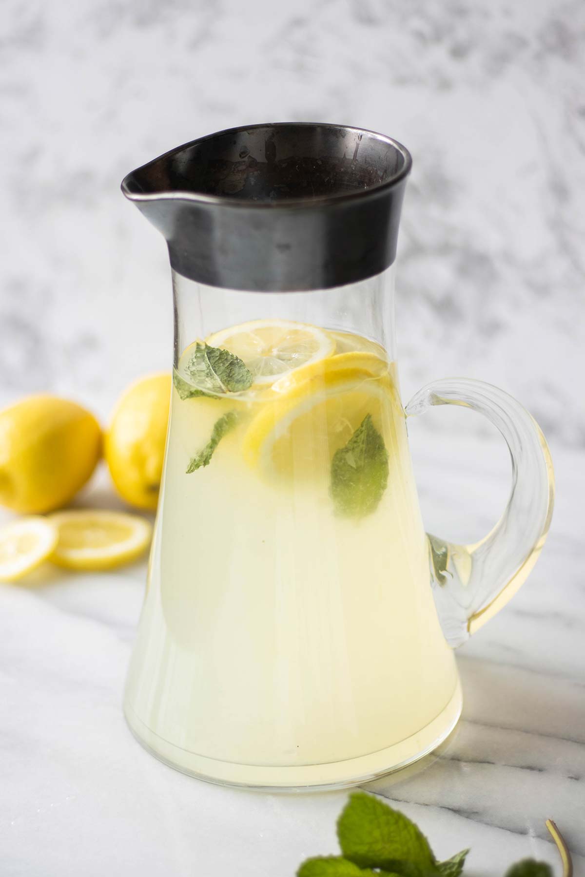 a big pitcher full of lemon mint juice with some lemons in the background and mint in the front.