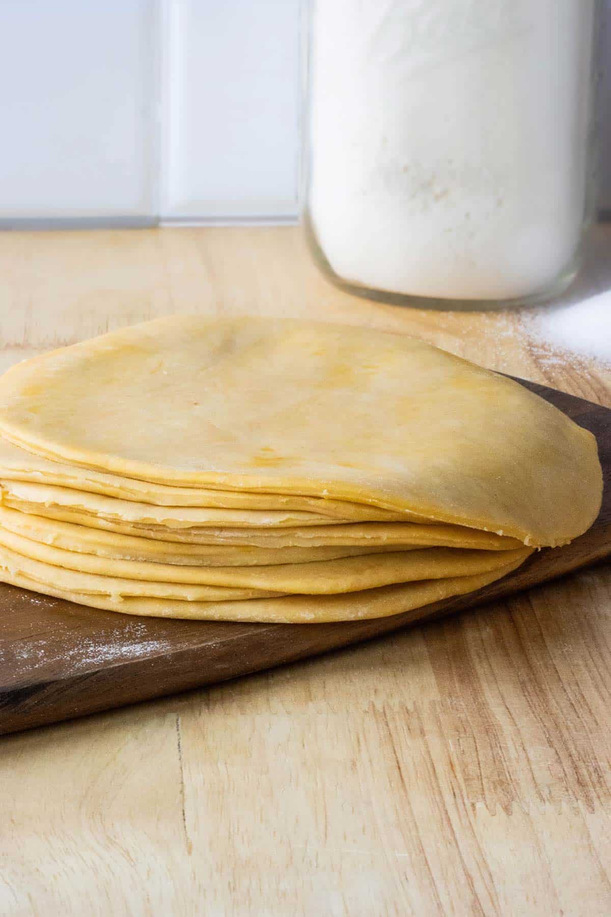 discos de masa de empanadillas sobre cortador de madera oscuro con un envase grande de harina en el fondo y un poco de sal sobre la mesa.