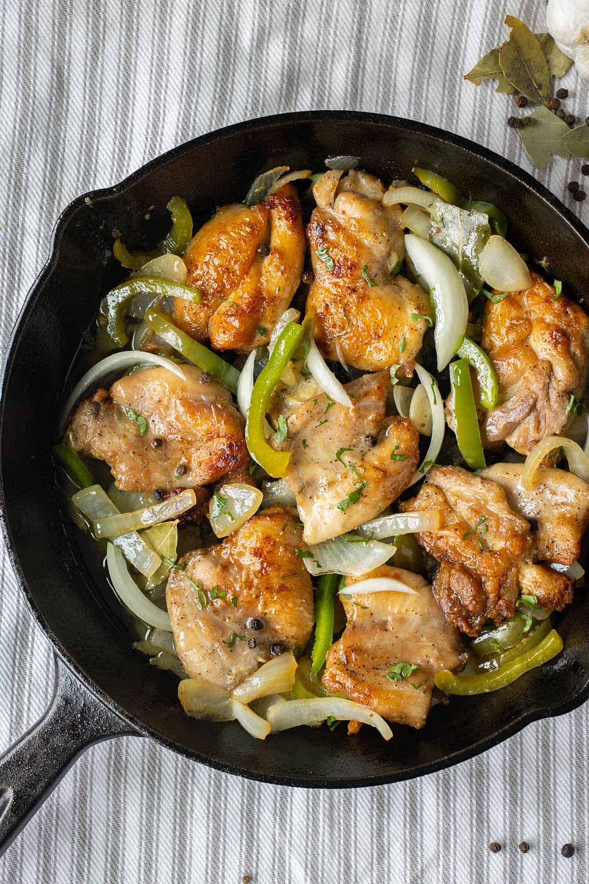 sarten de hierro negro con un pollo en escabeche sobre mantel de rayas blanco y gris con varias hojas de laurel y pimienta a un lado.