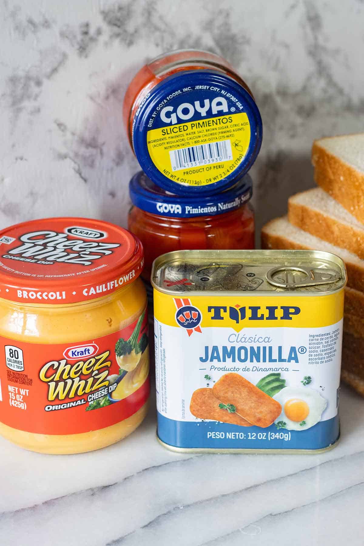 a jar of cheeze whiz, 2 jars of goya sliced pimientos, tulip brand luncheon meat and a few slices of bread on a stack.