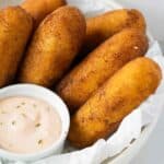 a white plate with parchment paper and 6 sorullos de maiz on top with a small dish with mayoketchup sauce on the side.