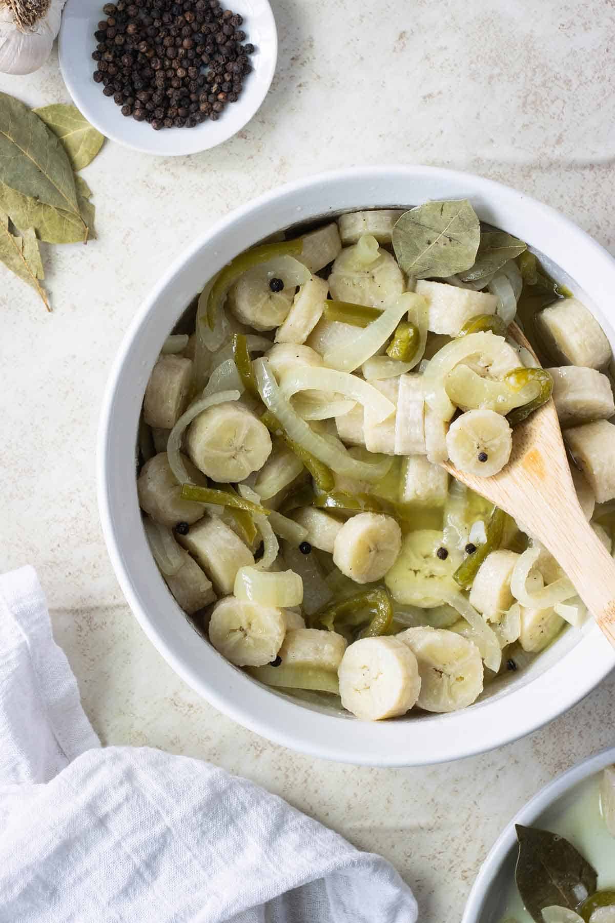 a big white bowl with guineos and escabeche and a wooden spoon with some garlic, black pepper and bay leaves as garnish.