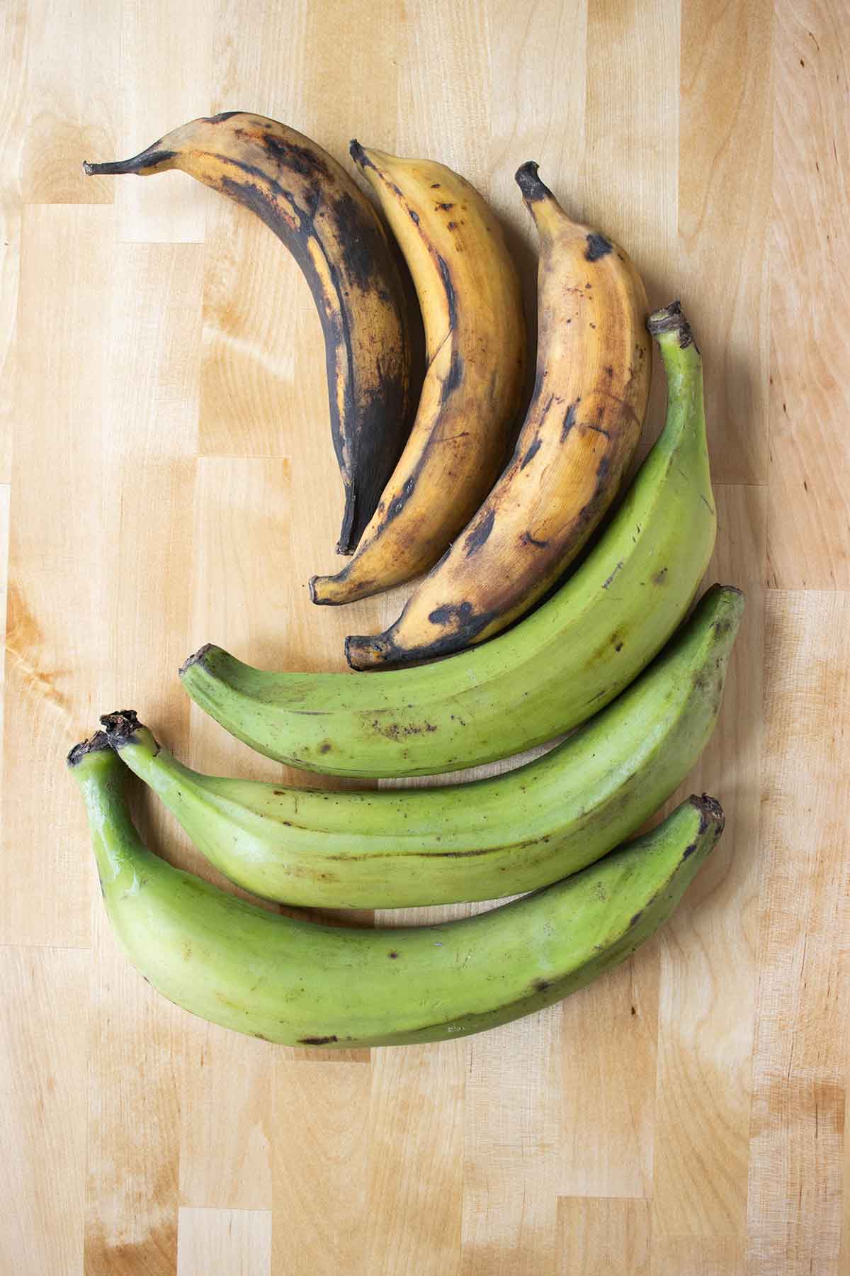 6 plantains on a table, 3 ripe plantains next to 3 green plantains.