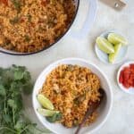 a pan and a plate with arroz con jueyes with different plates with tostones, limes and red peepper and a beer and cilantro on the side.
