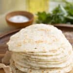 a stack of fresh yuca tortillas over a wood cutting board with a salt dish of salt, parsley and oil on the background.
