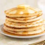 a plate with a stack of 5 coconut pancakes with syrup and a piece of butter on top with a couple of forks on the side.