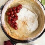a coconut milk dutch baby garnished with a cherry filling with some powder sugar and mint leaf on the side.