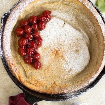 a coconut milk dutch baby garnished with a cherry filling with some powder sugar and mint leaf on the side.