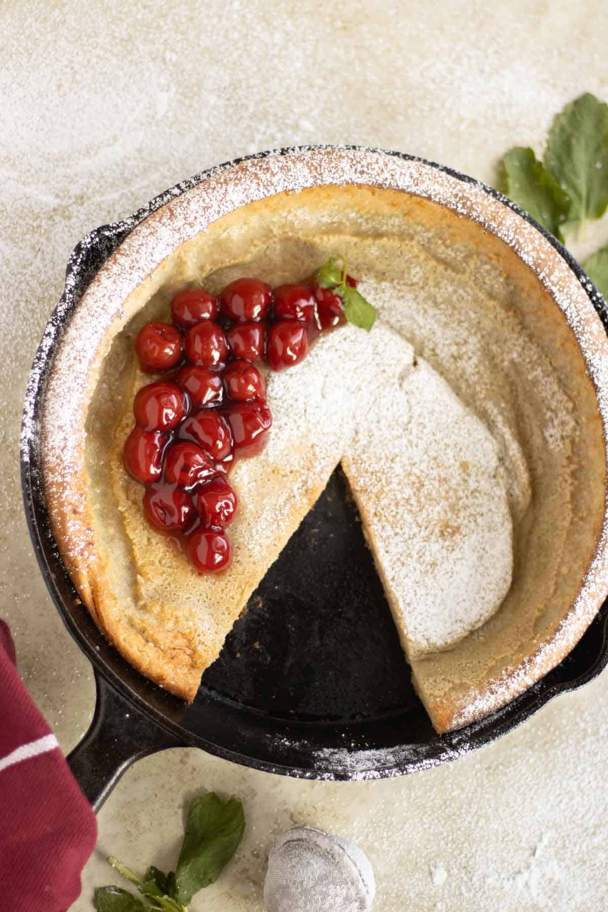 un sarten con un pancake holandes que tiene cherries como topping y azucar blanca con varias hojas de menta alrededor.
