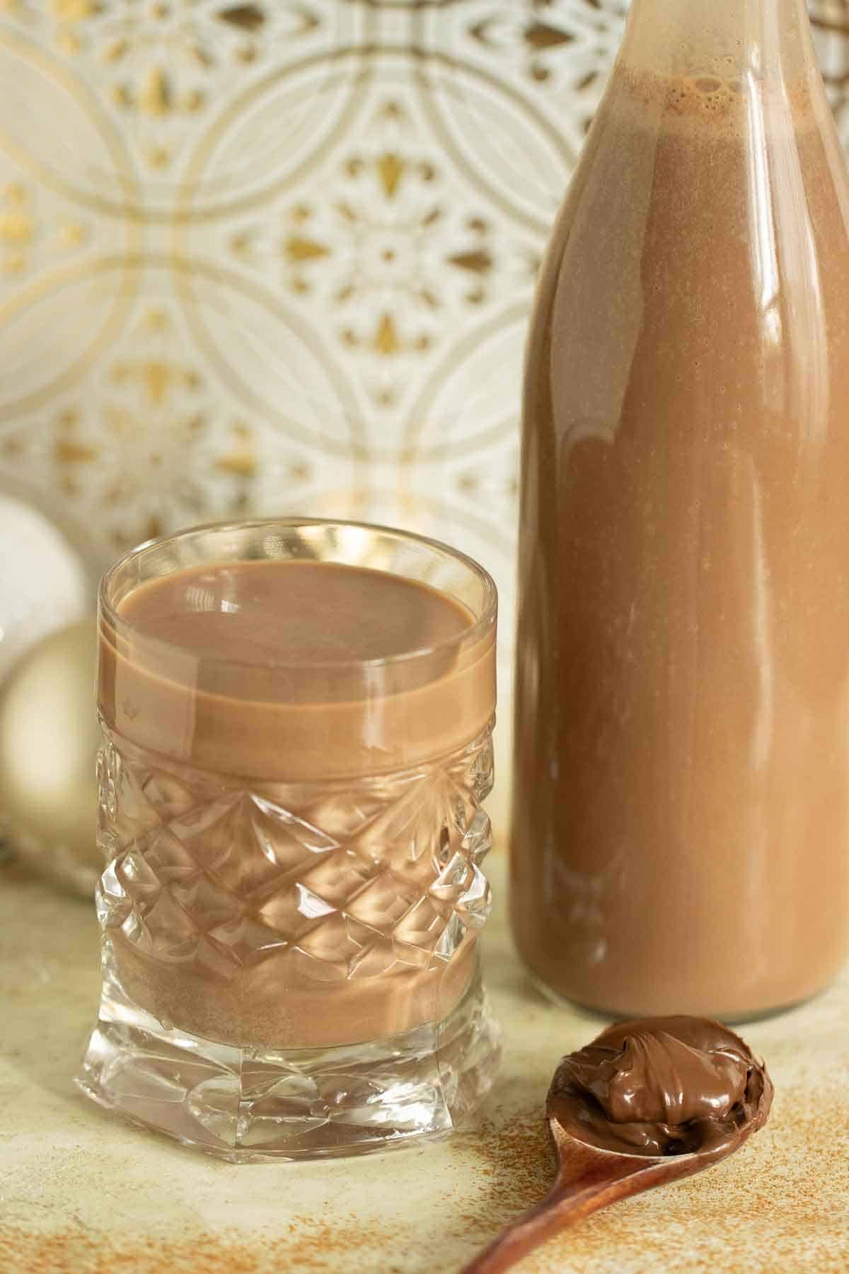 una taza llena de coquito de nutella con una botella llena detras y una cuchara con nutella al lado.
