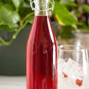 A bottle of a homemade strawberry syrup with a cup on the side.
