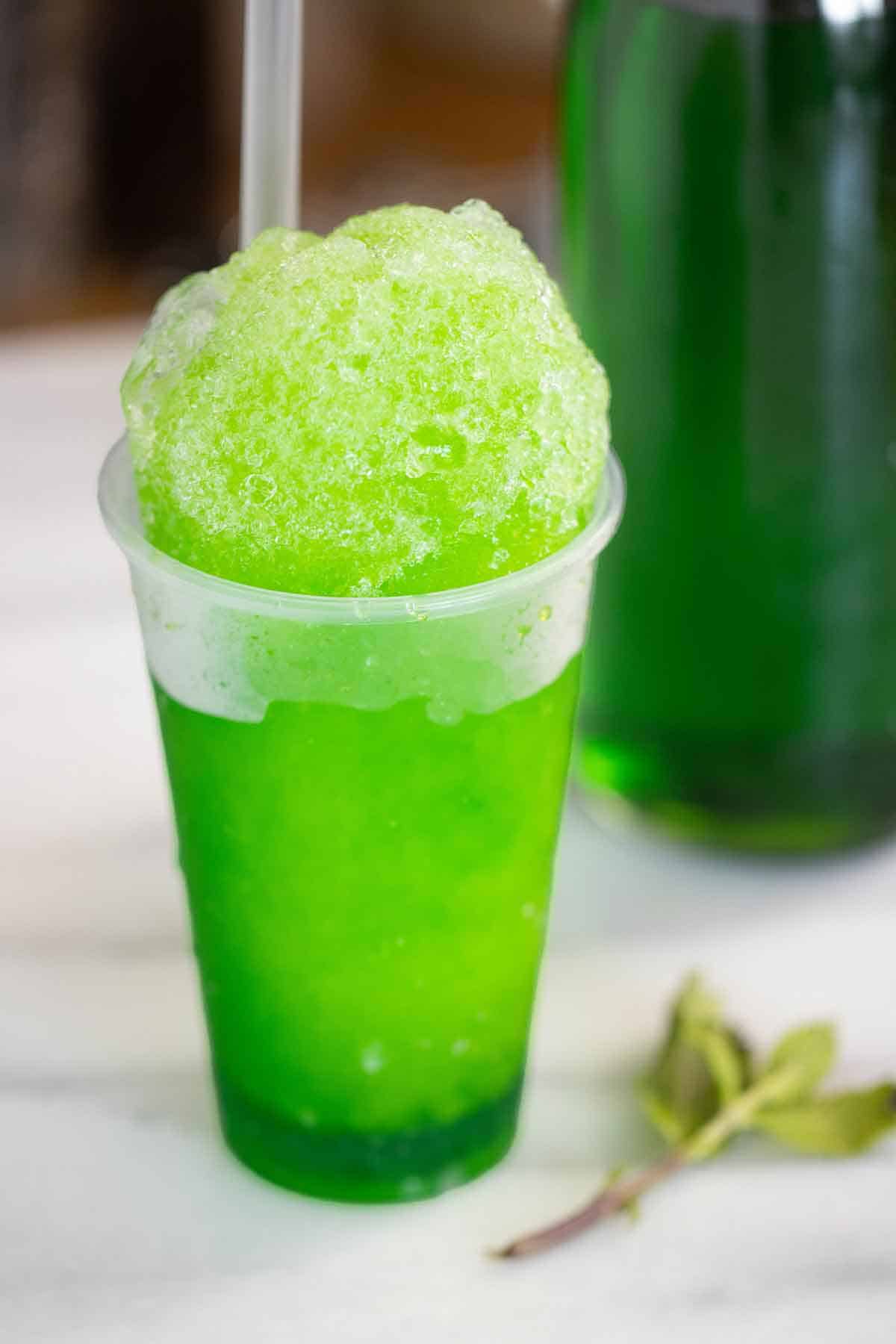 A mint flavor piragua with a fresh mint on the side and a syrup bottle on the background.