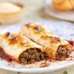A plate with 2 beef cannelloni with a garlic bread and parmesan cheese on the background.