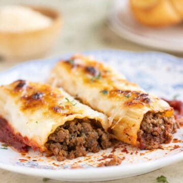 A plate with 2 beef cannelloni with a garlic bread and parmesan cheese on the background.