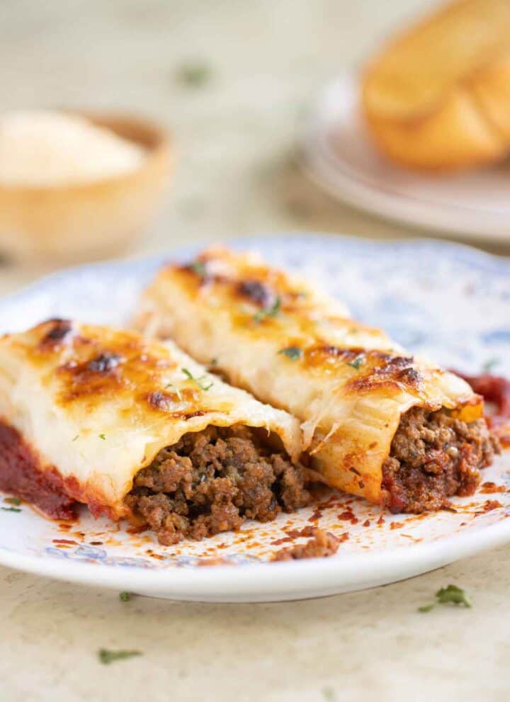A plate with 2 beef cannelloni with a garlic bread and parmesan cheese on the background.