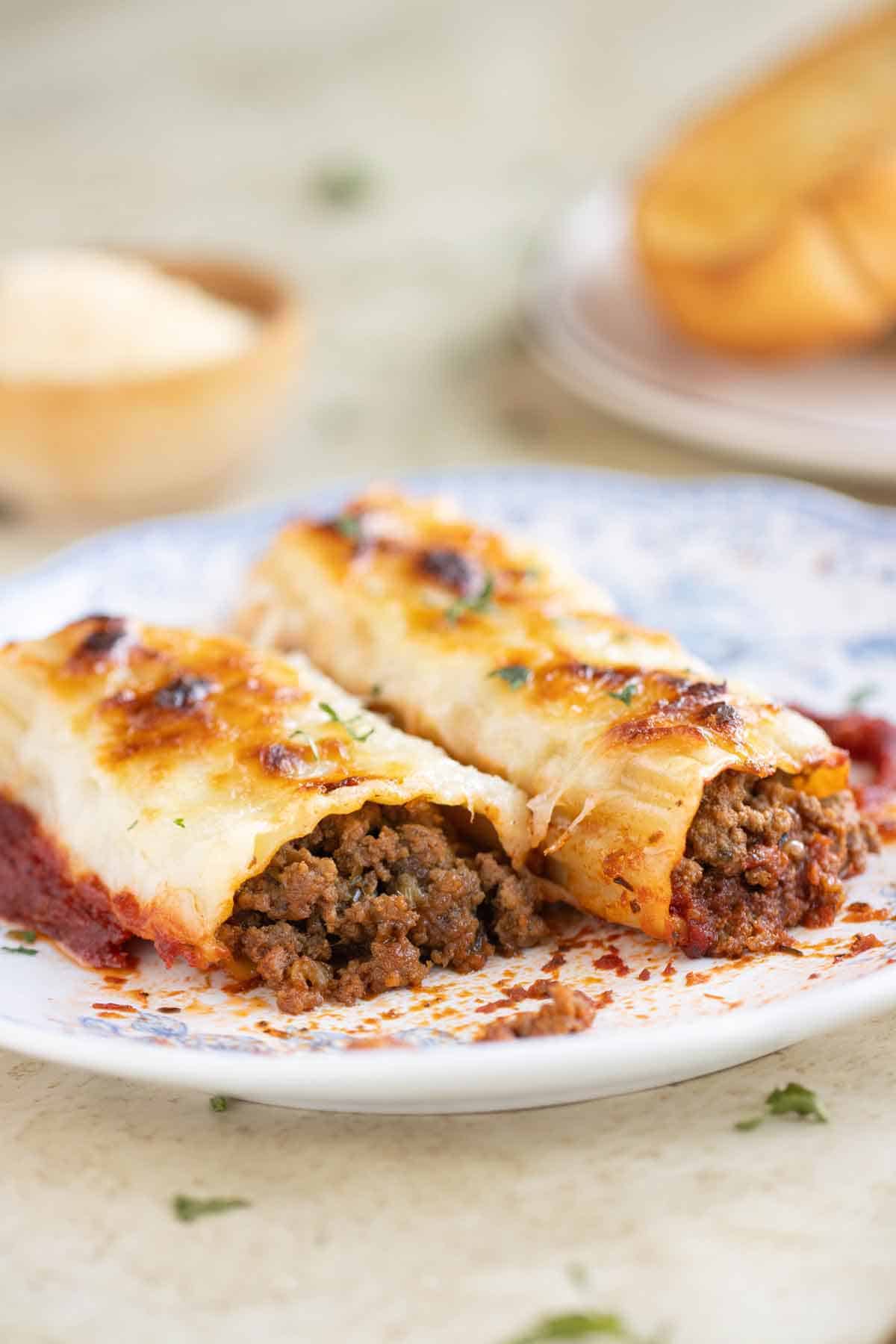 A plate with 2 beef cannelloni with a garlic bread and parmesan cheese on the background.