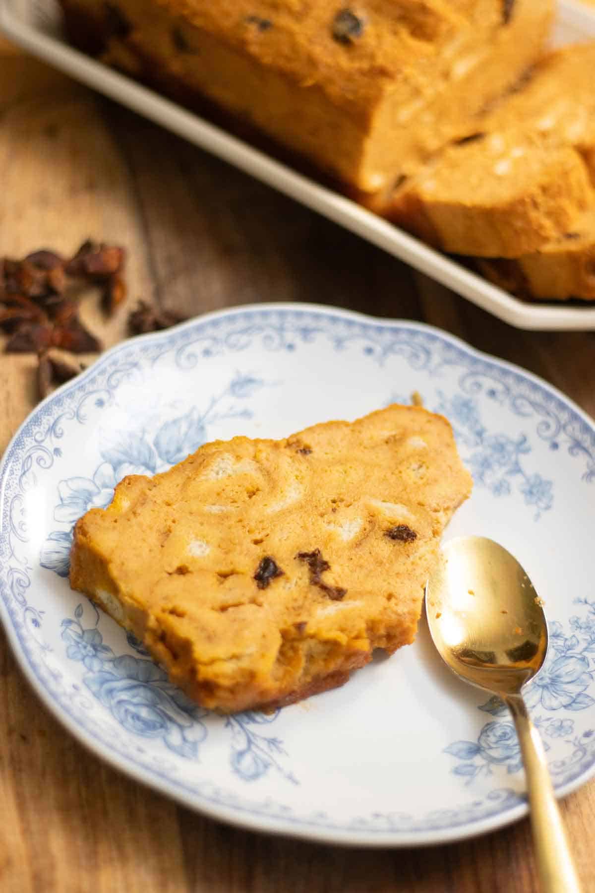A plate with a pumpkin bread pudding slice with a spoon and a big loaf of the bread pudding on the background.