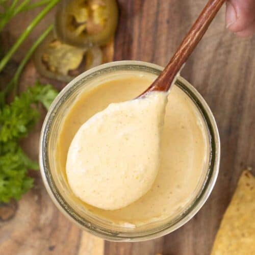 A. hand holding a spoon full of a creamy jalapeno sauce over a big jar with parsel, jalapeno pieces and, tostitos around.