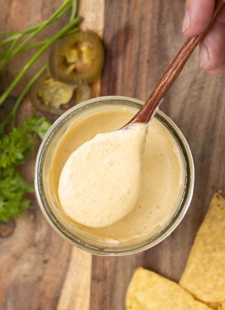 A. hand holding a spoon full of a creamy jalapeno sauce over a big jar with parsel, jalapeno pieces and, tostitos around.