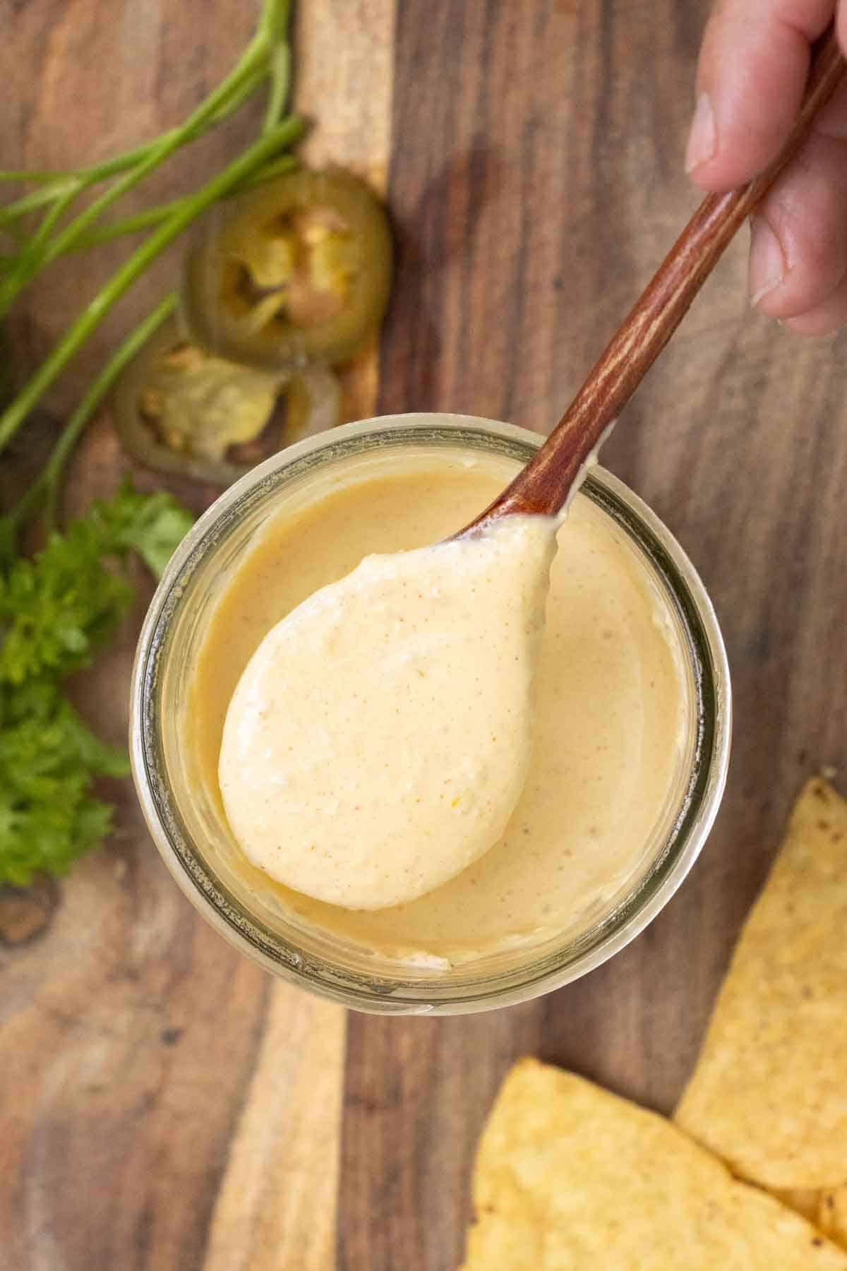 A hand holding a spoon full of a creamy jalapeno sauce over a big jar with parsley, jalapeno pieces and, tostitos around.