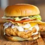 A fried chicken sandwich on a brioche bun with lettuce, tomato, corn tortilla chips, cheese, a creamy jalapeno sauce and onions.