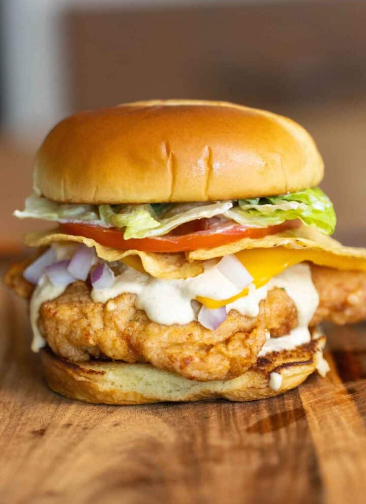 A fried chicken sandwich on a brioche bun with lettuce, tomato, corn tortilla chips, cheese, a creamy jalapeno sauce and onions.