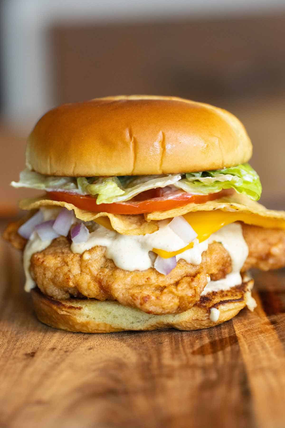 A fried chicken sandwich on a brioche bun with lettuce, tomato, corn tortilla chips, cheese, a creamy jalapeno sauce and onions.