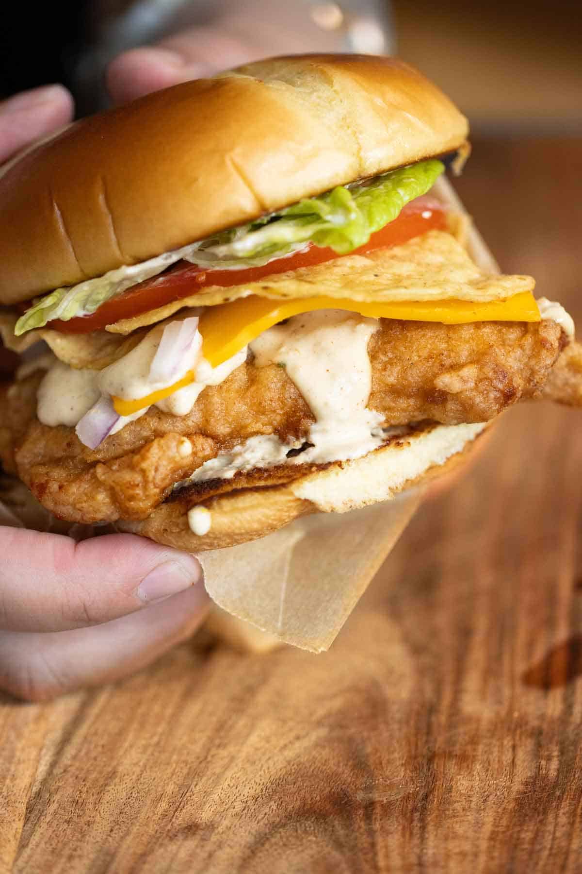 A pair of hands holding a crispy chicken sandwich made on a brioche burger bun with a creamy jalapeno sauce, cheese, tortilla chips, lettuce, tomato and onion.
