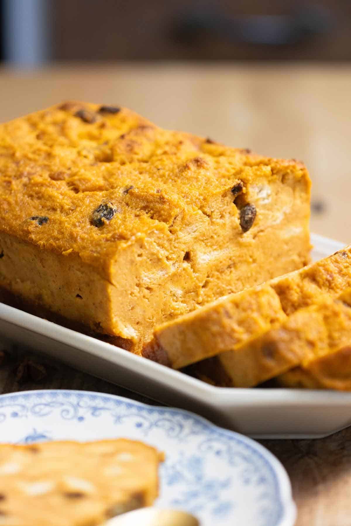 A big loaf of a pumpkin bread pudding with a few cut slices and a slice on a plate on the front.